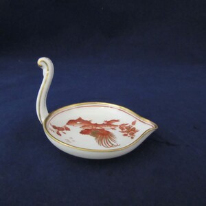 May include: A white porcelain spoon rest with a gold rim and a red and gold floral design. The spoon rest has a handle shaped like a swan's neck.
