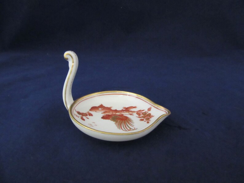May include: A white porcelain spoon rest with a gold rim and a red and gold floral design. The spoon rest has a handle shaped like a swan's neck.