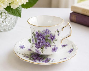 May include: A white porcelain teacup and saucer set featuring a delicate pattern of purple violet flowers and green leaves. The rim of both the cup and the saucer is accented with a thin gold trim, and the cup has a scalloped edge design.