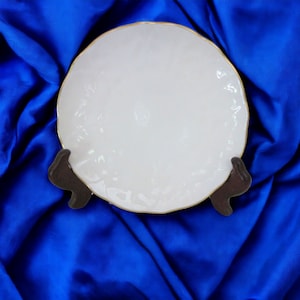 May include: A white, round plate with a textured surface and a gold-coloured rim. The plate is displayed on a dark brown stand against a backdrop of blue fabric.
