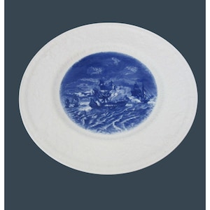 May include: White ceramic plate with a blue nautical scene in the center. The scene depicts ships at sea, possibly a naval battle. The plate's rim has a textured, embossed pattern.