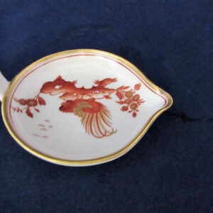 May include: A white porcelain gravy boat with a gold rim and a red and gold floral design. The boat has a long, curved handle.