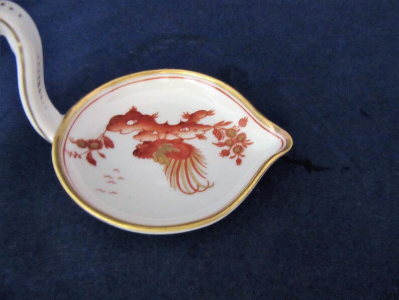 May include: A white porcelain gravy boat with a gold rim and a red and gold floral design. The boat has a long, curved handle.