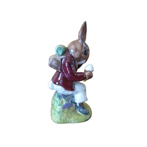 May include: A brown and white rabbit figurine wearing a red jacket and carrying a green and brown backpack. The rabbit is sitting on a green base with purple flowers.