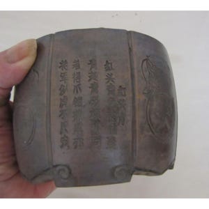 May include: A dark brown, cylindrical container with etched Chinese characters and decorative circular patterns. The container is held by a hand and appears to be made of a dense material. The text is partially visible.