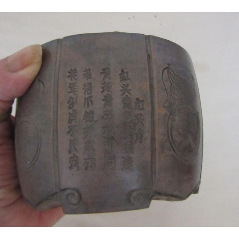 May include: A dark brown, cylindrical container with etched Chinese characters and decorative circular patterns. The container is held by a hand and appears to be made of a dense material. The text is partially visible.
