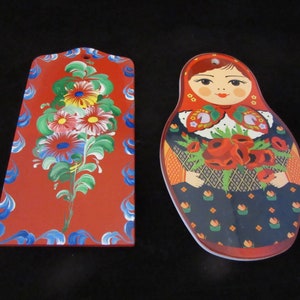 May include: Two wooden cutting boards with hand-painted designs. One board features a red background with a bouquet of flowers in blue, green, pink, and yellow. The other board is shaped like a Russian nesting doll and features a woman in a red and black dress holding a bouquet of red flowers.