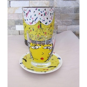 May include: A decorative glass cup, saucer, and cylindrical container. The cup and saucer feature a yellow face design with black outlines and red lips. The container has a yellow base with a white top and colorful confetti-like accents. The text "Mia Cara" is visible.