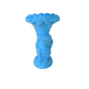 May include: A light blue glass vase featuring a ruffled, scalloped rim and a central sculptural figure of a seated person supporting the flared top. The base is circular and the entire object is rendered in a solid, opaque, bright blue color.