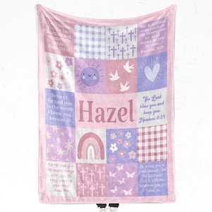 Könnte beinhalten: Eine rosa- und lavendelfarbene Patchwork-Decke mit dem Namen "Hazel" in der Mitte. Die Decke zeigt Blumen-, Kreuz- und Regenbogen-Designs sowie Bibelverse. Die Decke ist ein weiches, gemütliches Produkt.