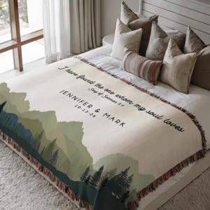 May include: A cream-coloured throw blanket featuring a mountain landscape design in shades of green and blue. The blanket has the text "I have found the one whom my soul loves" and the names "JENNIFER & MARK" with a date. The blanket is fringed.