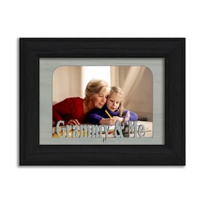 May include: A black picture frame with a silver mat and a photo of a grandmother and granddaughter. The photo shows the two sitting at a table, with the granddaughter drawing. The text "Grammy & Me" is cut out of the silver mat.
