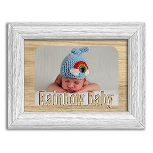 May include: A white wooden frame with a photo of a baby wearing a blue crocheted hat with a rainbow and clouds. The text "Rainbow Baby" is engraved on the frame.