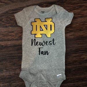 May include: Gray baby onesie with a gold Notre Dame logo and the text "Newest Fan" in black.