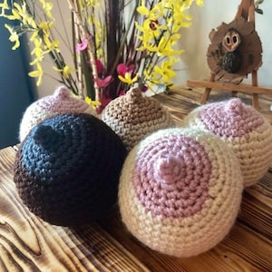 May include: Four crocheted breast shapes in different colors, including brown, pink, and beige. The shapes are arranged on a wooden surface.