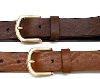 May include: Two leather belts arranged horizontally, one in a dark brown shade and one in a lighter tan brown. Each belt features a smooth, textured surface, a single leather keeper loop, and a polished gold-tone metal buckle with a rounded frame.
