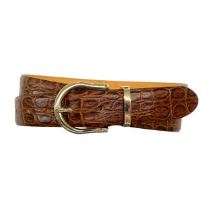 Puede incluir: Un cinturón marrón con un patrón texturizado en relieve de cocodrilo. El cinturón cuenta con una hebilla de metal dorado pulido y una trabilla de metal dorado a juego, mostrando un diseño clásico con un forro interior liso de color beige.