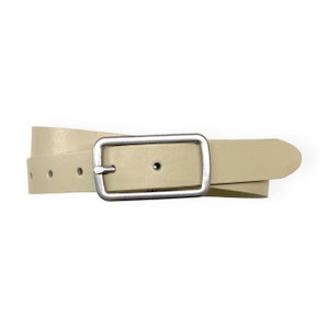 Può includere: Una cintura in pelle color beige chiaro con finitura liscia e una fibbia rettangolare in metallo color argento. Il cinturino presenta fori circolari distanziati uniformemente per la regolazione e una punta arrotondata.