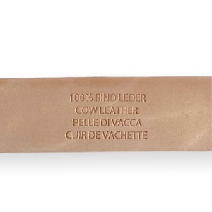 May include: A light tan leather strap featuring an embossed stamp that reads 100% RIND LEDER, COW LEATHER, PELLE DI VACCA, and CUIR DE VACHETTE. The surface of the leather is smooth and uniform in color, displaying the text clearly in a sans-serif font.