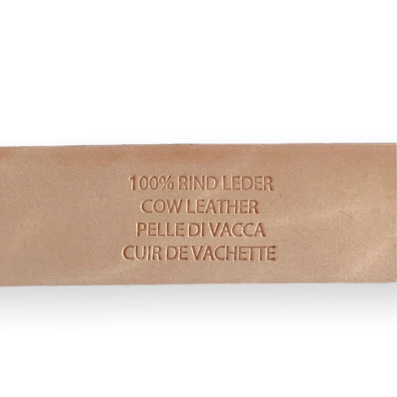 May include: A light tan leather strap featuring an embossed stamp that reads 100% RIND LEDER, COW LEATHER, PELLE DI VACCA, and CUIR DE VACHETTE. The surface of the leather is smooth and uniform in color, displaying the text clearly in a sans-serif font.