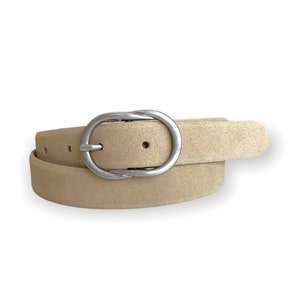 Op de afbeelding: Een beige riem met een suède-achtige textuur en een gladde, ovaalvormige gesp van zilverkleurig metaal. De riem heeft zichtbare verstelgaatjes en is in een cirkelvorm opgerold, wat de neutrale kleur en de metalen hardware goed laat zien.
