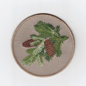 Puede incluir: Un parche bordado beige con una rama de pino verde y dos piñas marrones.