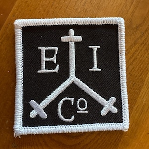 Puede incluir: Parche cuadrado negro con texto y diseño bordados en blanco. El diseño incluye una cruz, las letras "E" e "I", las letras "C" y "o", y dos espadas cruzadas. El parche tiene un borde blanco.