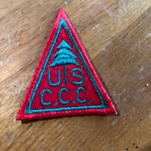 Może przedstawiać: Czerwona trójkątna naszywka z zieloną obwódką i akcentami. Naszywka posiada zielony wzór drzewa na górze oraz zielone litery "UIS C.C.C.". Naszywka jest prawdopodobnie zabytkowym emblematem lub odznaką.
