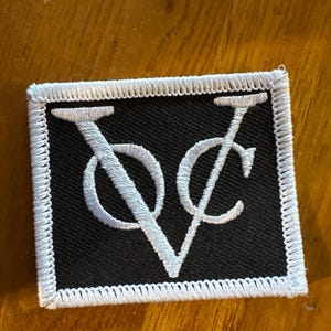 Op de afbeelding: Een rechthoekige patch met een zwarte achtergrond en witte geborduurde letters "V" en "C" die met elkaar verweven zijn. De patch heeft een witte rand met een gestikt ontwerp.