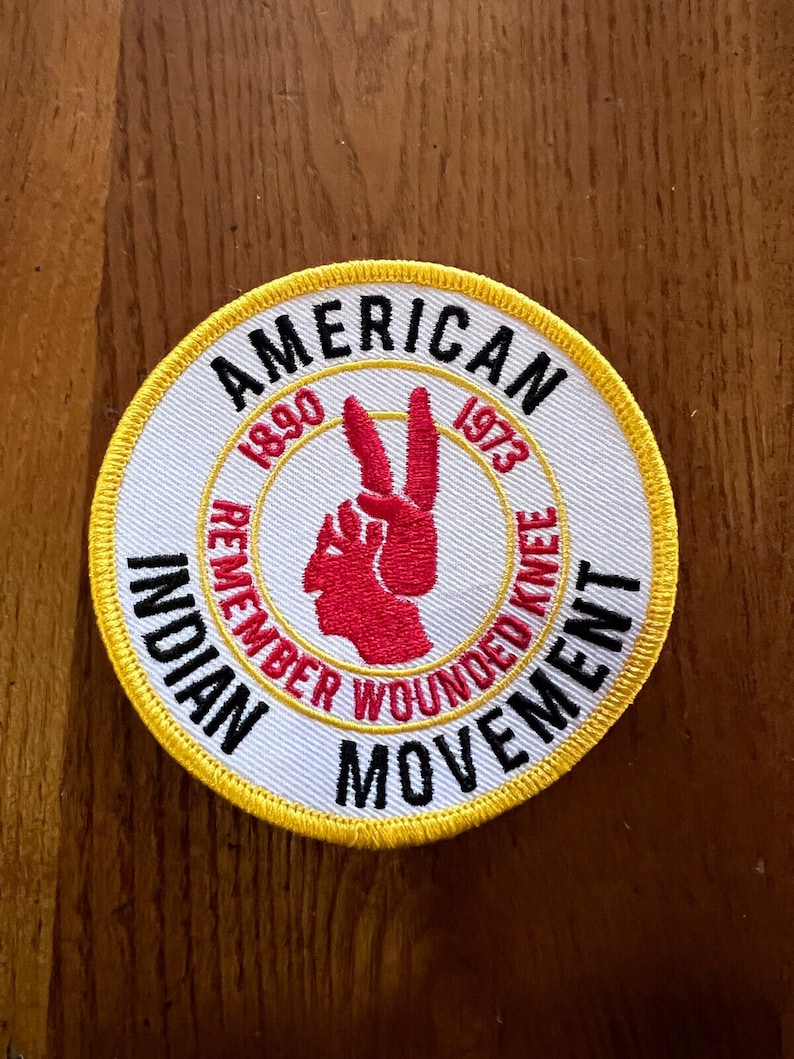 K&ouml;nnte beinhalten: Ein wei&szlig;er runder Patch mit gelbem Rand. Der Patch hat schwarzen Text, der "American Indian Movement" und "Remember Wounded Knee 1890 1973" lautet. Ein rotes Peace-Zeichen mit einem roten Profil eines amerikanischen Ureinwohners befindet sich in der Mitte des Patches.