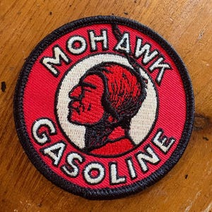 May include: Round, embroidered patch with the text "MOHAWK GASOLINE" in white lettering on a red background. The patch features a profile of a Native American man in red and black, set against a white circle, with a black border.