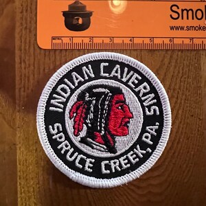 Könnte beinhalten: Ein runder weißer Patch mit schwarzen und weißen Stickereien. Der Patch zeigt ein Profil eines amerikanischen Ureinwohners mit roten und weißen Federn und dem Text "INDIAN CAVERNS SPRUCE CREEK, PA."