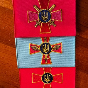以下が含まれることがあります： 金色の十字架に、円の中に青い三叉槍が描かれた刺繍パッチが3つ。 上のパッチは赤、真ん中のパッチは薄い青、下のパッチは赤です。