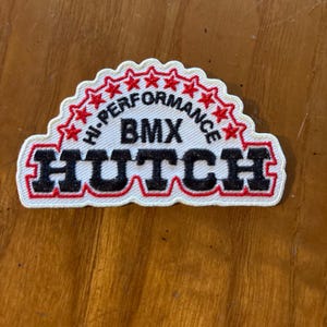 Peut inclure: Patch brodé blanc avec des coutures rouges et noires. Le patch présente le texte "HI-PERFORMANCE BMX HUTCH" avec une rangée d'étoiles rouges au-dessus du texte.