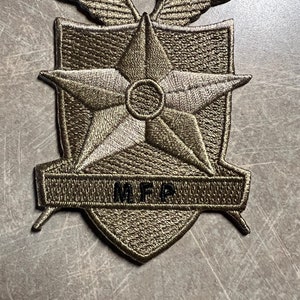 Op de afbeelding: Een groene geborduurde patch met een vijfpuntige ster, vleugels en de tekst "M.F.A." in zwart.