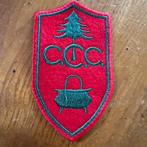 May include: A red shield-shaped patch with green trim. The patch features a green pine tree, the letters "C.C.C.", and a green cooking pot. The patch is likely a vintage emblem or insignia.