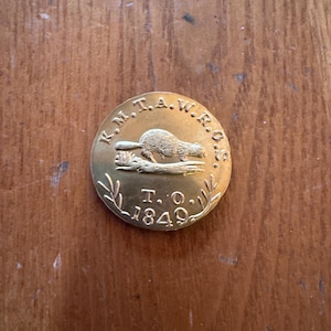 May include: A circular, gold-toned metal badge featuring embossed text and a beaver illustration. The text around the edge reads "K.M.T.A.W.R.O.S." with "T.O. 1849" below the beaver. The badge is set against a wooden surface.