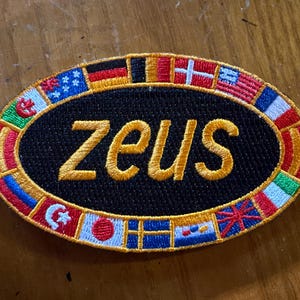 May include: Embroidered patch with the word "zeus" in gold thread on a black oval. The border features a ring of colorful flags. The patch is likely for a jacket or bag.