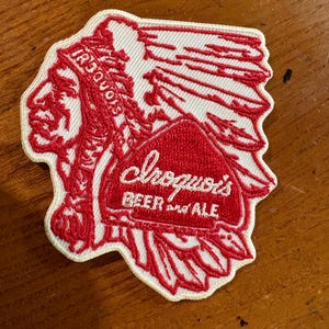May include: Embroidered patch featuring a Native American chief in red and white. The patch has the text "Iroquois" and "Iroquois Beer and Ale" in a stylized font. The patch is on a white background with a red border.