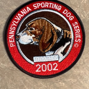 May include: A red and black embroidered patch with a white border. The patch features a brown and white Beagle dog with the text "Pennsylvania Sporting Dog Series" and "2002" in white.