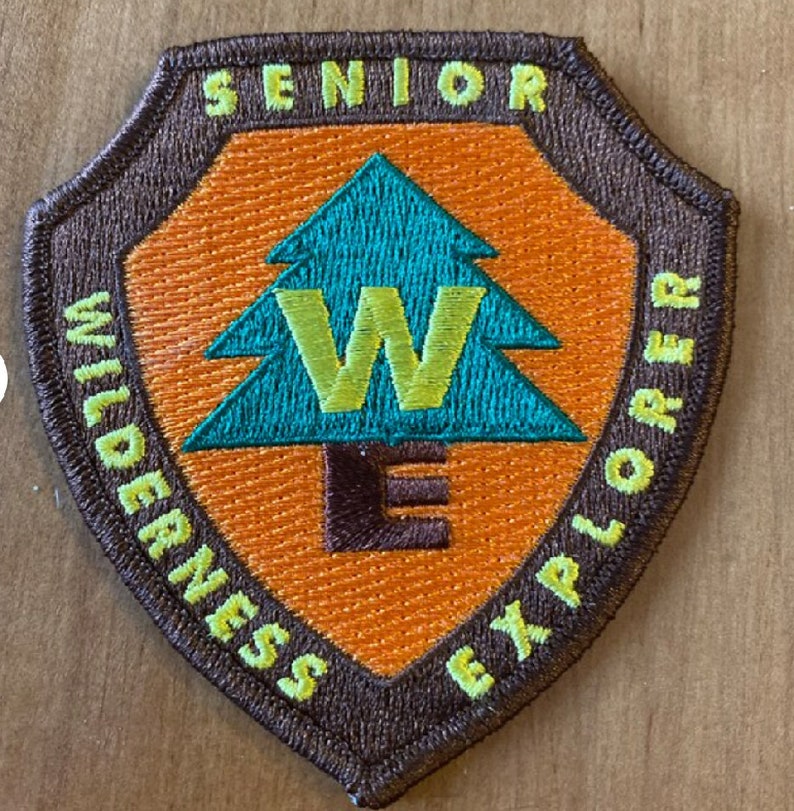 May include: A brown, green, and yellow embroidered patch with the text "Senior Wilderness Explorer" and a stylized green pine tree with a yellow "W" inside.