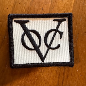 May include: Rectangular patch with a white background and a black border. The patch features the intertwined letters "V" and "C" in black, creating a stylized design. The patch is likely an emblem or logo.