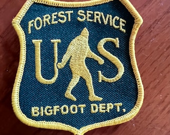 May include: A dark green patch with a yellow border, depicting a stylized Bigfoot walking. The patch reads "FOREST SERVICE US BIGFOOT DEPT."