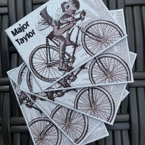 May include: Five white embroidered patches with a brown image of a person riding a bicycle. The text "Major Taylor" is on the top patch.