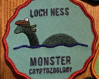 May include: A teal patch with a red scalloped border features the text "LOCH NESS MONSTER CRYPTOZOOLOGY." The patch depicts the Loch Ness Monster wearing a hat, emerging from blue water. The patch is a fun, whimsical design.