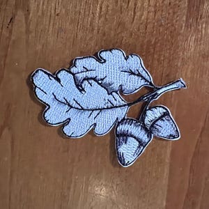 Puede incluir: Un parche bordado plateado de una hoja de roble con dos bellotas. La hoja de roble está detallada con venas y las bellotas están sombreadas.