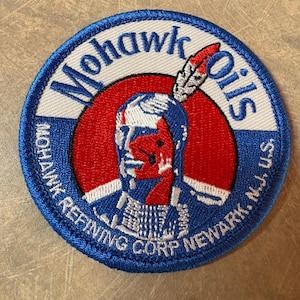 Peut inclure: Un écusson circulaire avec une bordure bleue et les mots "Mohawk Oils" en blanc. Le centre représente une figure amérindienne en rouge, blanc et bleu, avec une coiffe de plumes. Le texte "Mohawk Refining Corp Newark, N.J. U.S." est également présent.