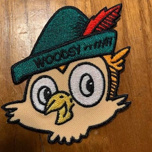 May include: A green and red embroidered patch of a cartoon owl wearing a green hat with a red feather. The hat has the text "Woodsy" on it.