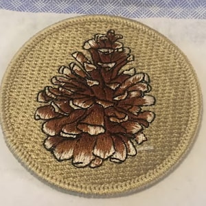 Pode incluir: Um pinhão bordado de cor castanho sobre um fundo de tecido bege. O pinhão é detalhado com pontos de costura brancos.