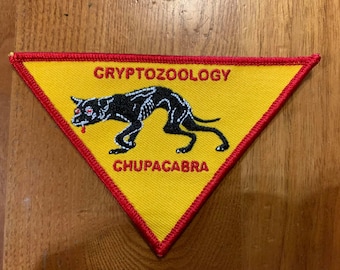 May include: A yellow triangular patch with a red border. The patch features a black silhouette of a chupacabra with red eyes and a dripping mouth. The text "CRYPTOZOOLOGY" is above the creature and "CHUPACABRA" is below.